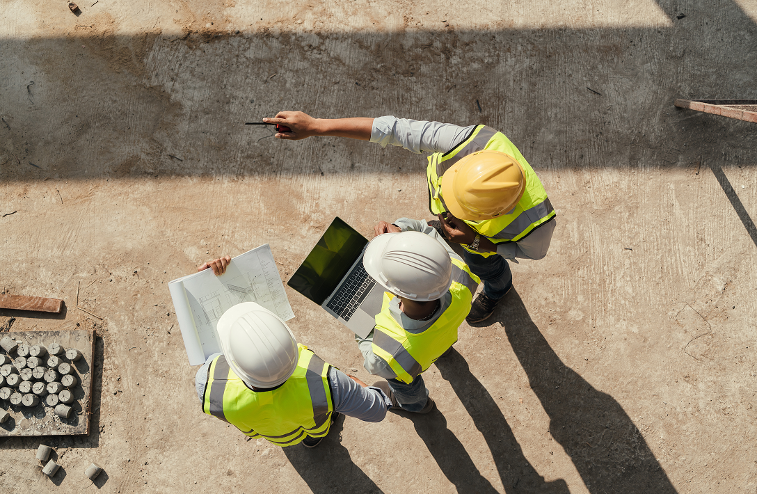 Image of three construction workers on site working off plans and a laptop. Image taken from overhead